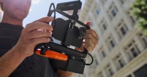 A person holds a professional video rig with a mounted smartphone and microphone setup, possibly for filming. In the background, there is a tall building and some greenery, indicating an outdoor urban setting.