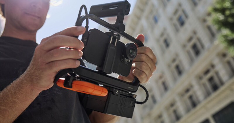 A person holds a professional video rig with a mounted smartphone and microphone setup, possibly for filming. In the background, there is a tall building and some greenery, indicating an outdoor urban setting.