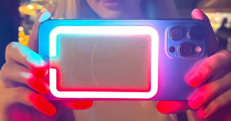 A person holds a smartphone with a glowing rectangular attachment on the back, lit up in blue and red colors. The smartphone's dual camera lenses are visible, and part of the person’s face can be seen in the background.