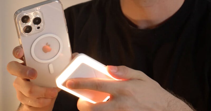 A person holding a smartphone with a prominent Apple logo and a camera setup in one hand while attaching or using a small, lit rectangular device on the back of the phone with the other hand. The person is wearing a black shirt.