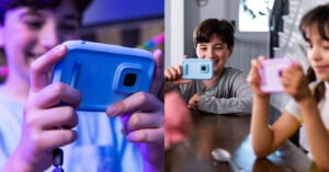 Two children are holding and looking at their own blue and purple handheld gaming devices. The child on the left is shown in a close-up view, smiling brightly. The child on the right is also smiling while sitting at a table with another child.