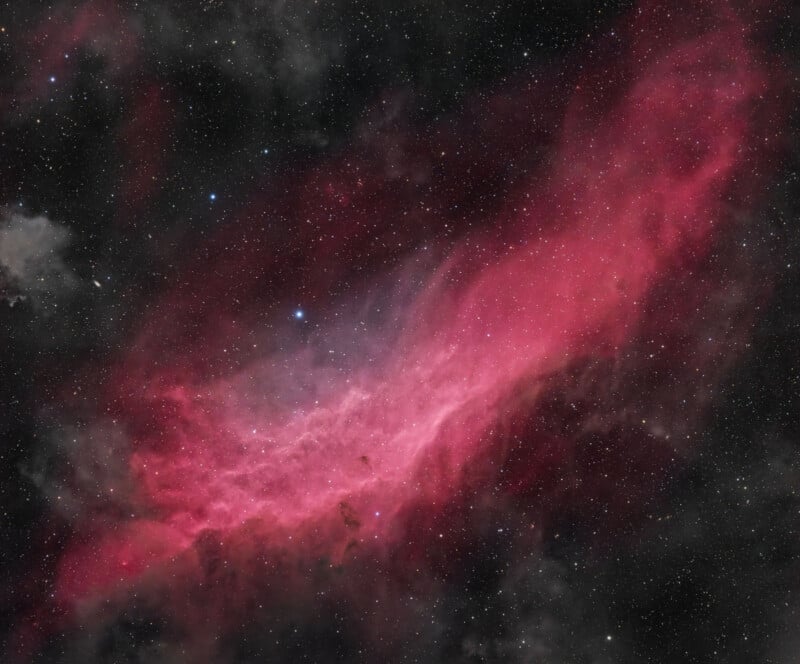 A colorful nebula with vivid pink and red hues set against a dark backdrop of space. Multiple bright stars are scattered throughout the image, adding contrast to the cloudy, glowing gas and dust formation. The nebula appears to be expansive and ethereal.