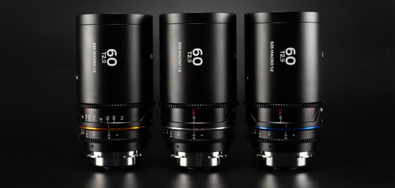 A close-up of three black camera lenses in a row, each labeled "60 2x 85mm micro." The lenses have metallic bands near the base in three different colors: gold on the left, white in the center, and blue on the right. The background is dark.