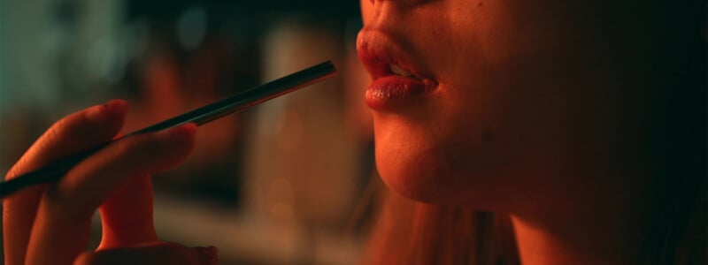 A close-up shot of a person's lips and fingers holding a straw near their mouth. The background is out of focus, creating a soft, warm, and intimate atmosphere with dim lighting. The person's lower face is bathed in a gentle, warm light.