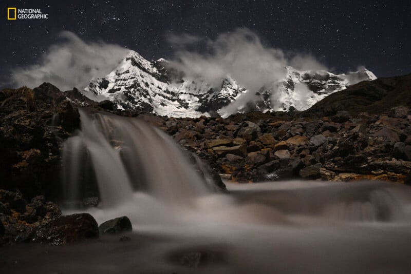 A nighttime mountain scene with snow-covered peaks beneath a starry sky. A smooth, blurred waterfall cascades over rocks in the foreground. The National Geographic logo is in the top left corner.