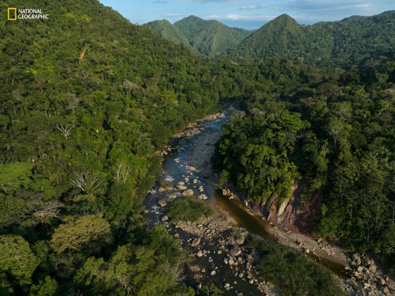 Aerial view of a dense, tropical forest with a rocky river flowing through it. The area is lush with green vegetation, surrounded by rolling hills and mountains in the background. The image includes a National Geographic logo in the top left corner.