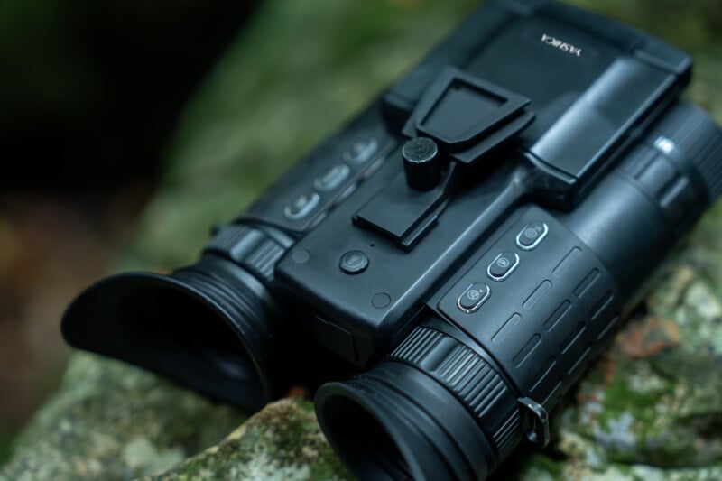 A pair of black binoculars with rubber grips rests on a rock. The binoculars feature multiple buttons and a viewing screen on top, suggesting advanced functionalities. The background shows an out-of-focus natural environment.