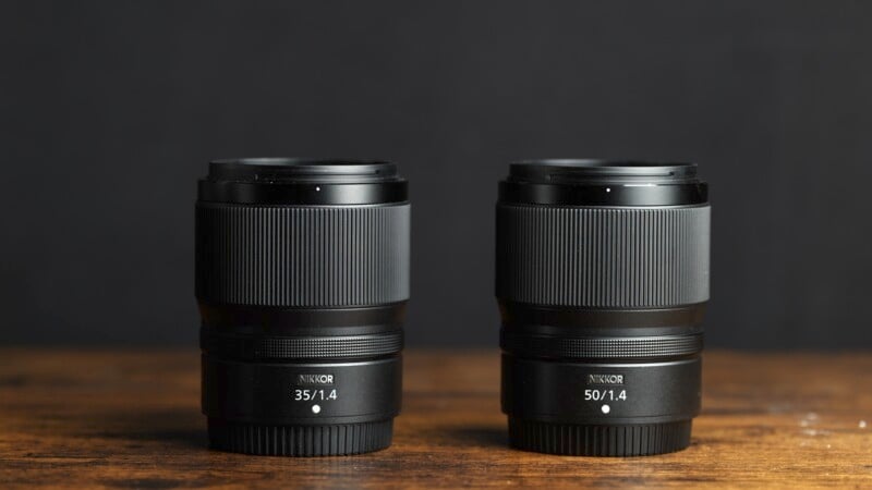 A photograph of two NIKKOR camera lenses on a wooden surface with a dark background. The lens on the left is labeled "35/1.4," and the one on the right is labeled "50/1.4." Both lenses are black with a similar design.