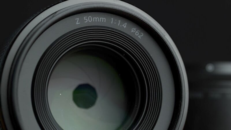 Close-up shot of a camera lens with inscriptions "Z 50mm 1:1.4 Ø62" around the front element against a dark background. The lens glass reflects light, revealing internal aperture blades.