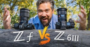 A man enthusiastically poses behind two cameras placed on a log with their lenses facing forward. The cameras are labeled "Z f" and "Z 6III". The text "Z f" and "Z 6III" is prominently displayed in front of the cameras with a "VS" symbol separating them. The background features greenery.