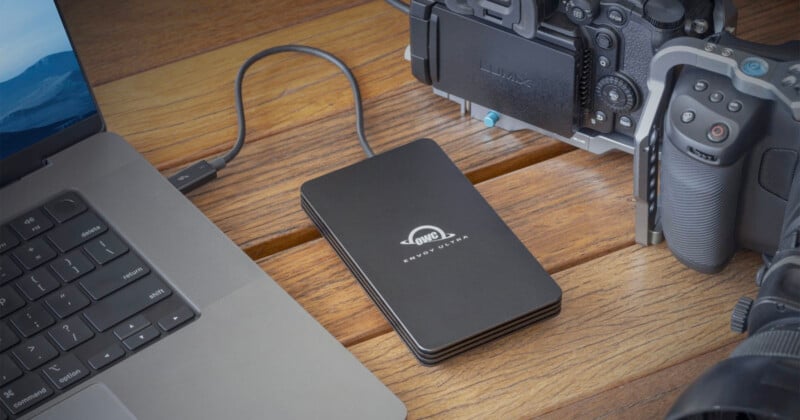 A black external hard drive, labeled "OWC Envoy Pro Elektron," connected to a laptop via a USB-C cable, placed on a wooden surface. A digital camera is partially visible next to the hard drive.