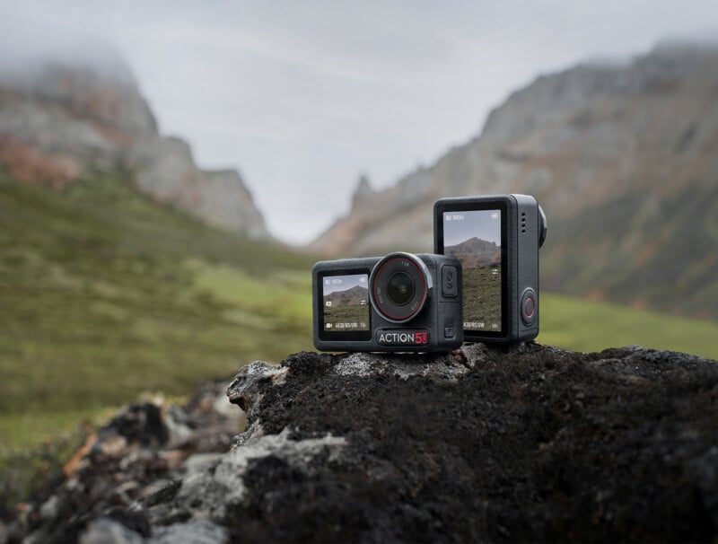 Two action cameras are placed on a rock with a scenic mountainous landscape in the background. One camera is standing upright, showing its screen displaying the same landscape, while the other camera lies flat. Both cameras have "Action 3" labeled on them.