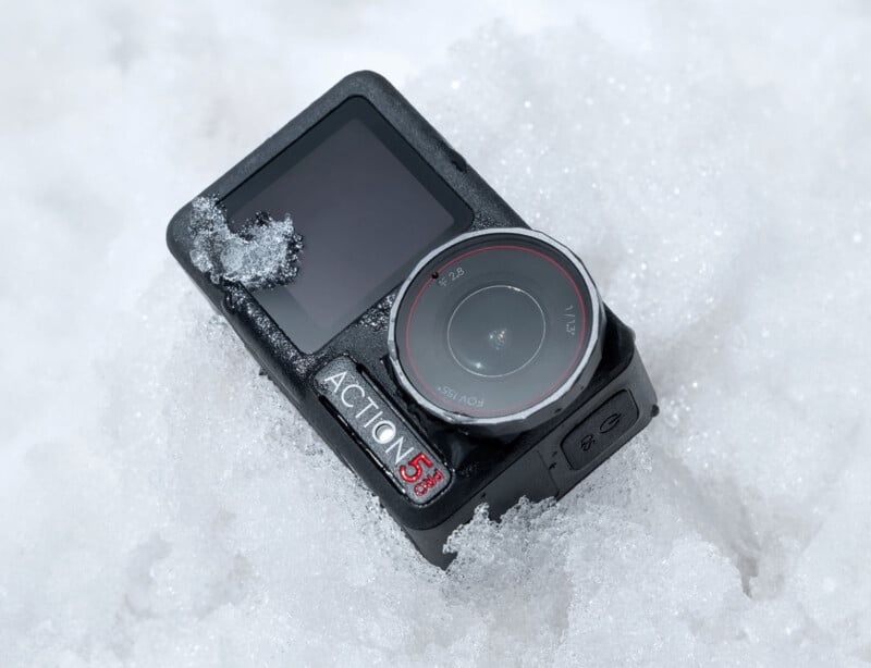 A black Action 5S camera partially covered in snow, lying on a snowy surface. The camera lens has a ring with "F2.8" and "POV 47" markings, and the screen appears to be off. Snow is visibly accumulated on the lens and body of the camera.