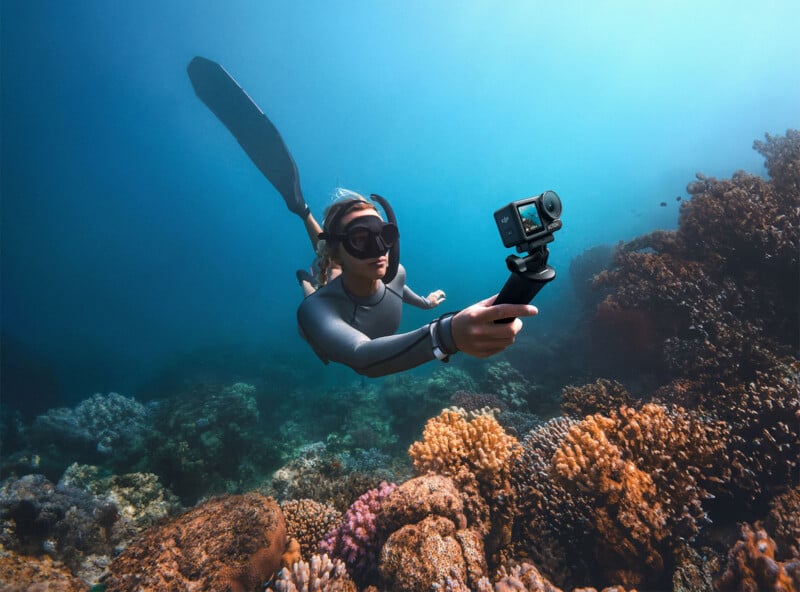 A person in a wetsuit and snorkeling gear is diving underwater, holding a camera on a selfie stick, surrounded by vibrant coral reefs of various colors and shapes in clear blue water.