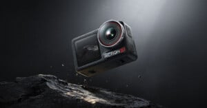 A rugged black action camera labeled "ACTION" with a lens and display is suspended mid-air above a rocky surface, illuminated by a directed light from above. Dust particles float around it, highlighting its robust and adventure-ready design.