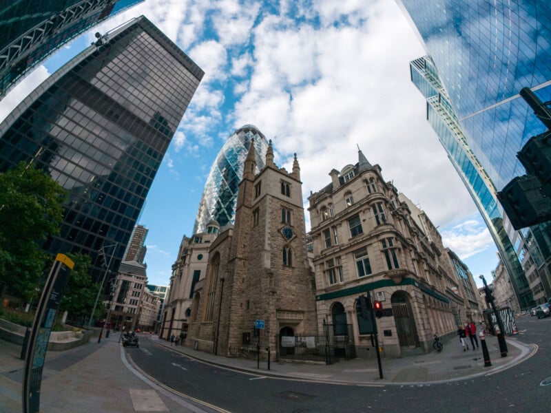 A street scene in London with historic and modern skyscrapers, featuring the iconic Gherkin building, a stone church, and other contemporary office buildings under a partly cloudy sky. The view is captured with a fisheye lens, creating a curved distortion.