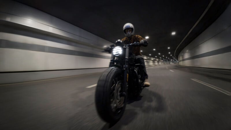 A motorcyclist rides through a dimly lit tunnel at high speed. The rider is wearing a helmet, and the motorcycle's headlights are on, illuminating the road ahead. The tunnel walls curve in the distance, giving a dynamic sense of motion.