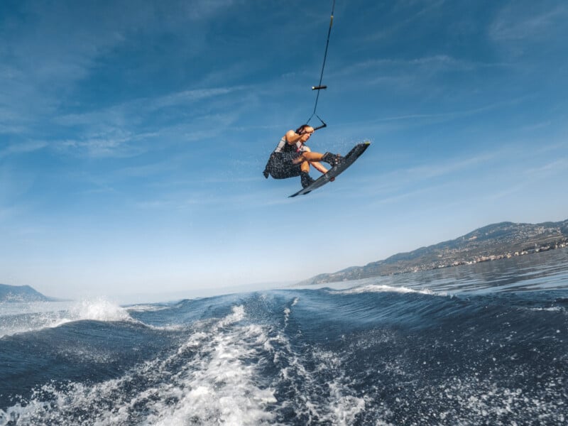 A person is wakeboarding on a body of water, performing an aerial trick while holding onto a tow rope. The sky is clear, and mountains are visible in the background, with water splashing behind the wakeboarder.