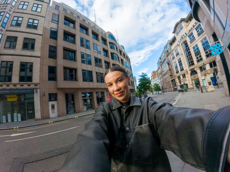 A woman wearing a black jacket takes a selfie on a city street with modern buildings in the background. The sky is partly cloudy, and the street appears quiet with no visible traffic or pedestrians.