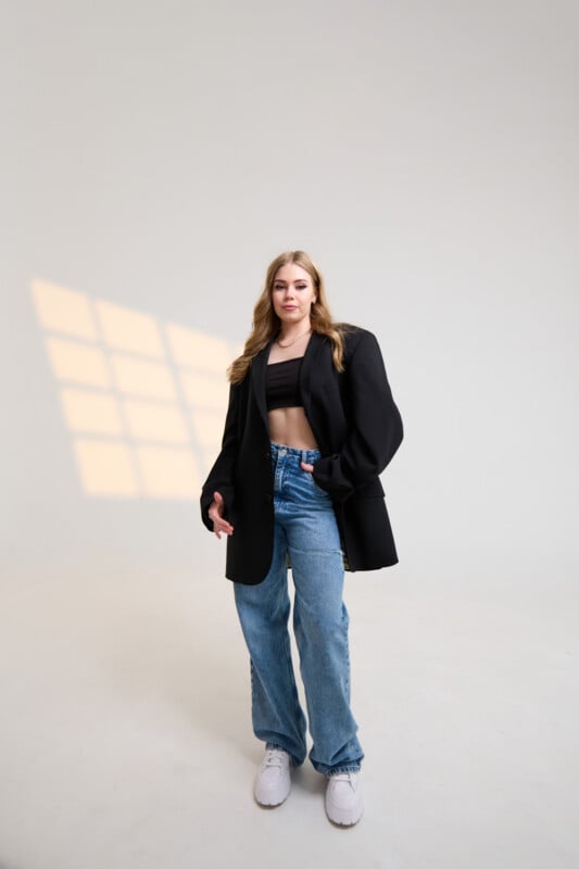 A woman standing in a minimalist room with a window casting a grid pattern of sunlight on the wall. She is wearing a black blazer over a black crop top, blue high-waisted jeans, and white sneakers. She has long, light brown hair and is looking confidently at the camera.