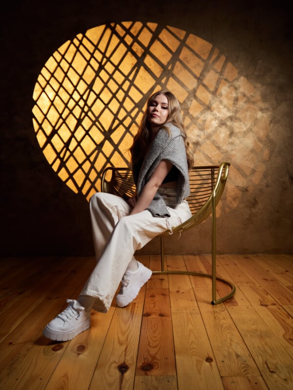 A person with long hair sits on a gold chair in front of an illuminated, circular lattice pattern on a brown textured wall. They are dressed in light-colored pants, white chunky sneakers, and a gray top, with a relaxed and contemplative expression. The floor is wooden.