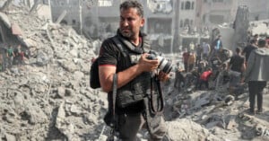 A man wearing a press vest and holding a camera looks to his left while standing on rubble. The background shows a chaotic scene with destroyed buildings and people searching through the debris. The atmosphere is dusty and tense.