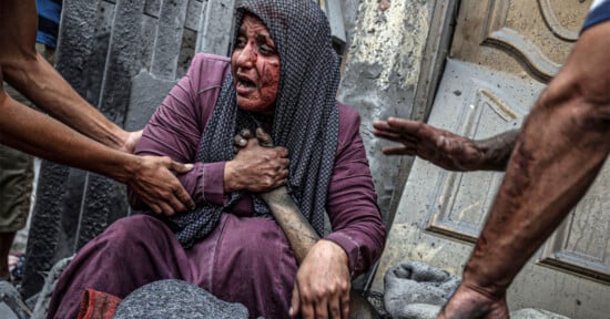 A distressed, bloodied woman in a headscarf clutches her chest, surrounded by reaching hands and rubble. Her face is marked by grief and pain, indicating a recent traumatic event in a chaotic environment.