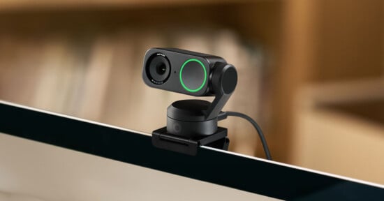 A compact black webcam with an integrated microphone and green LED indicator is mounted on the top edge of a computer monitor. The background shows blurred shelves with books.