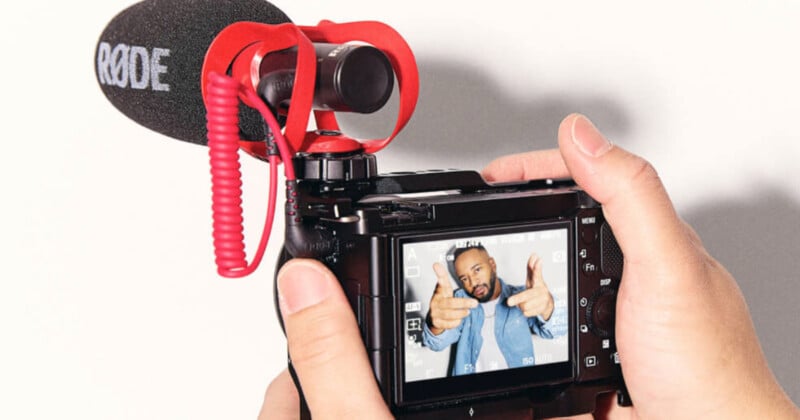 A person holds a camera with a large microphone attached, pointing it towards the viewer. The camera's screen displays an image of the same person pointing finger guns towards the lens, wearing a light blue jacket.