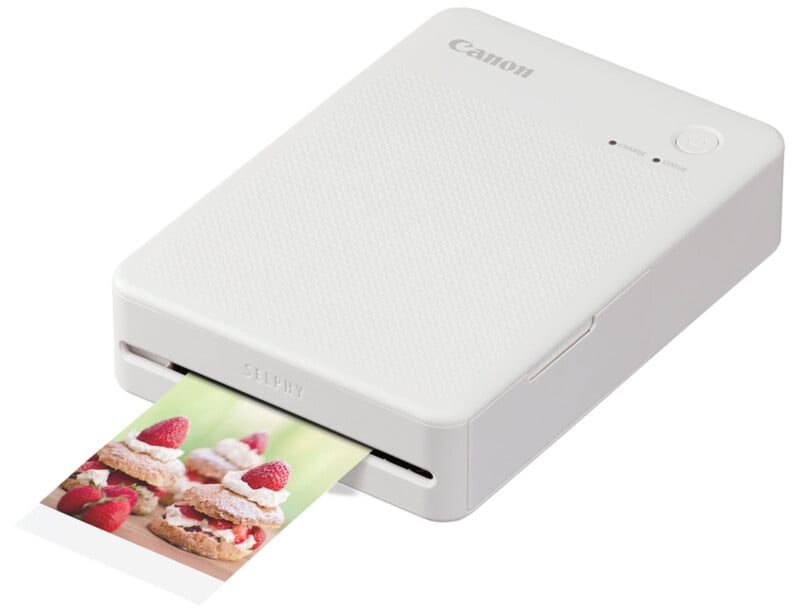 A compact white Canon Selphy photo printer is printing a photo of delicious cupcakes topped with raspberries and cream.