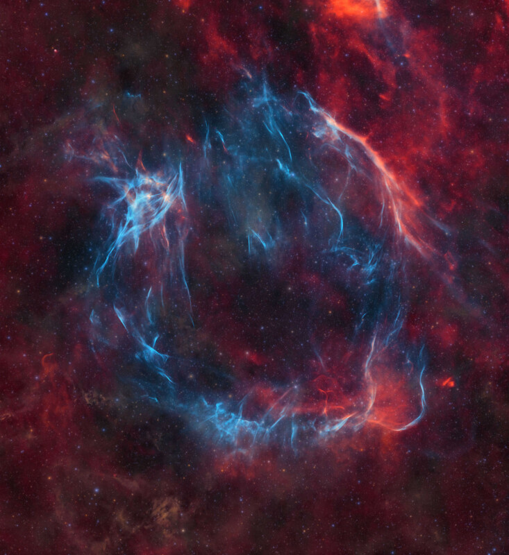 A stunning image of a cosmic scene features a vivid ring-like supernova remnant. Bright blue strands of gas and dust intertwine with fiery red and orange hues, creating a mesmerizing, ethereal display against a dark star-filled background.