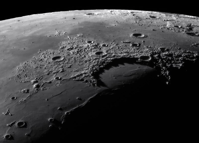 Close-up of the lunar surface showing a detailed view of numerous craters. A large shadow-filled crater is prominently visible, casting dark silhouettes against the rugged, cratered terrain of the Moon. The image presents a stark and intriguing lunar landscape.