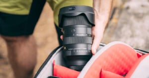 A person is holding a Sigma camera lens above an open camera bag with a red interior. The lens is black with various markings and a lens hood attached. The scene takes place outdoors, as indicated by the background.