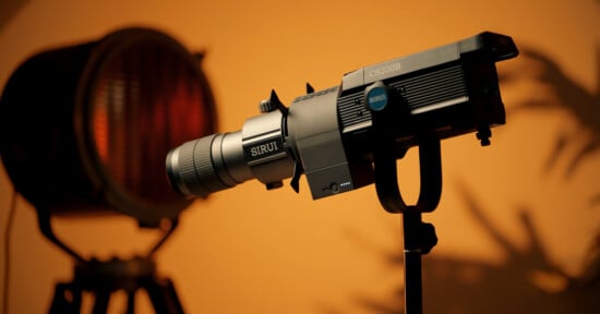 A professional camera with the brand name "SIRUI" is mounted on a tripod, facing left. It is set up in a studio with warm, ambient lighting, and a blurred background featuring a large spotlight. Shadows of plants can be seen on the wall.