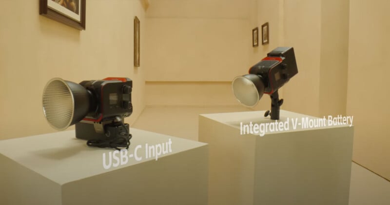 Two studio lights are displayed on separate pedestals in a minimalist room. One light has the text "USB-C Input" next to it, and the other has "Integrated V-Mount Battery" written beside it. The background features framed photos on the walls.