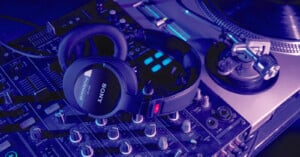 A pair of black Sony professional headphones rests on a DJ mixing console illuminated in blue and purple lighting. To the right, an old-fashioned turntable is partially visible, adding to the music production setting.