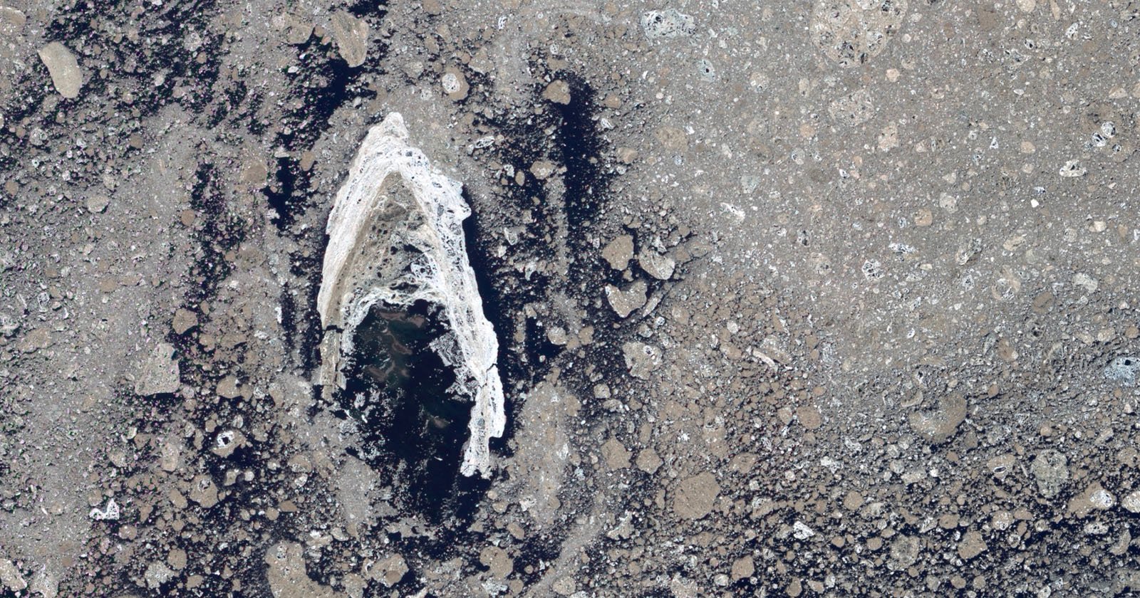 Aerial view of a barren, rocky landscape with a central elongated, white ice formation surrounded by darker terrain. The rugged surface is dotted with various-sized rocks and patches of black, creating a stark contrast with the icy feature.