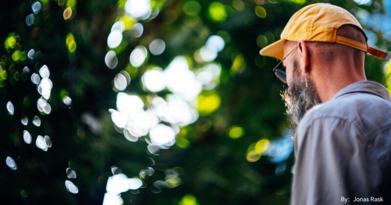 A bald man with a grey beard, wearing a yellow cap and sunglasses, is seen from the back. He stands in front of a blurred green and white bokeh background, likely an outdoor natural setting. (By: Jonas Rask).