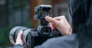 A person adjusts the settings on top of a professional DSLR camera. The camera has a large zoom lens attached, and the person's fingers are manipulating a button or dial near the viewfinder. The background is blurred, focusing attention on the camera.