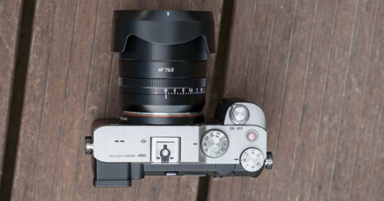 Top view of a digital camera on a wooden surface. The camera has a large attached lens with "AF 75/2" marked on it. Various control dials, buttons, and labels like "SteadyShot INSIDE" and "4K" are visible on the camera's body.