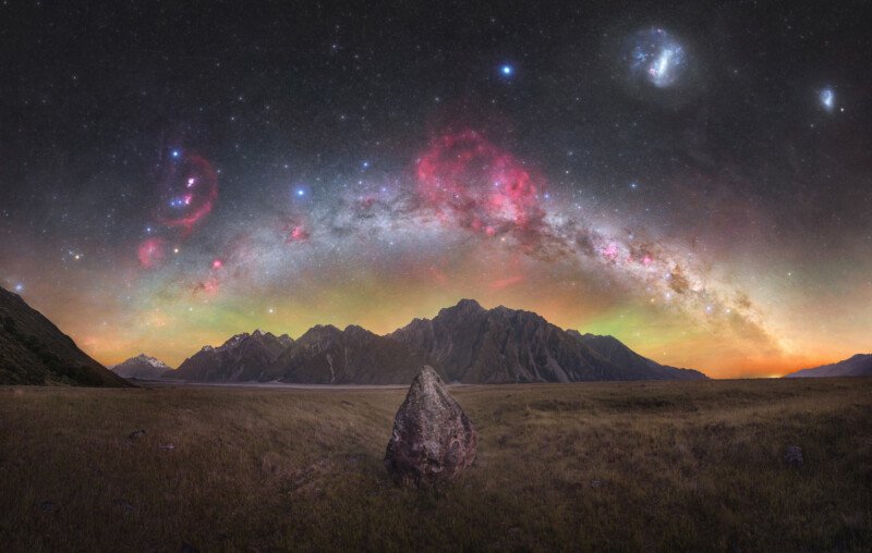 A vast night sky illuminated with bright stars and colorful nebulae arches over a mountainous landscape. In the foreground, a solitary large rock sits in a grassy field. Snow-capped peaks and rugged cliffs rise in the distance under the vibrant sky.