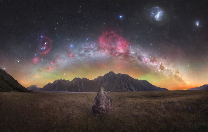 A vast night sky illuminated with bright stars and colorful nebulae arches over a mountainous landscape. In the foreground, a solitary large rock sits in a grassy field. Snow-capped peaks and rugged cliffs rise in the distance under the vibrant sky.