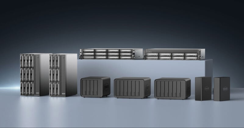 The image depicts various server equipment arranged in a row. There are large silver server racks, two rack-mounted servers on a platform, several black network-attached storage (NAS) units, and two black external hard drives, all on a gray surface against a dark background.