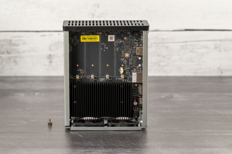 An exposed view of a small, rectangular computer case showing internal components including a motherboard, cooling fans, and circuitry. The case is open, with a small screw placed on the table next to it. The background is a textured gray surface.