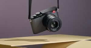 A black Leica camera with a textured body is suspended by its strap against a neutral background. The camera features a large lens with visible markings. It is positioned above abstract, geometric shapes in shades of beige and gold.