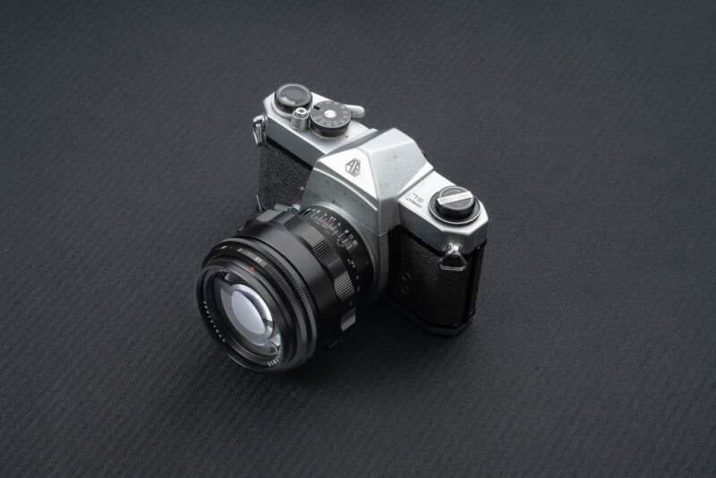 A vintage SLR camera with a silver body and a black textured grip is displayed on a dark, textured surface. The lens is prominently protruding from the front, featuring various adjustment dials and markings. The camera exudes a classic, retro appearance.