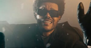 A person with short curly hair and a beard is wearing black sunglasses, a dark coat, a chain necklace, and black gloves. They have an expressive facial expression and are raising their hands. The lighting is moody with a hazy background.