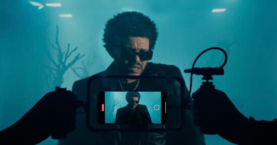 A person with curly hair, wearing sunglasses and a dark jacket, stands in a foggy, blue-hued setting. They are being filmed through a camera, which is visible in the foreground, held by two hands. Leafless branches can be seen in the background.