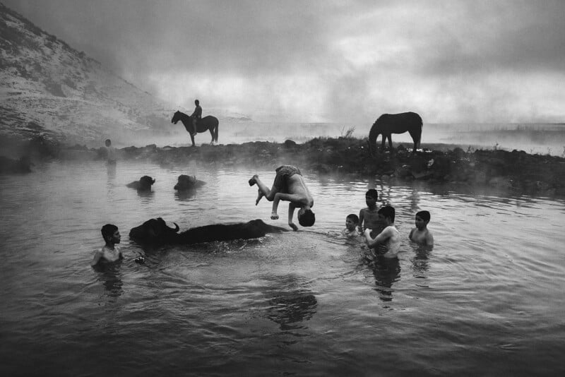 In a misty, mountainous landscape, several children play in a natural hot spring. One child flips off a submerged buffalo amidst the water. Two horses stand on the shore, one with a rider. The scene captures a moment of joy in a rugged, serene setting.