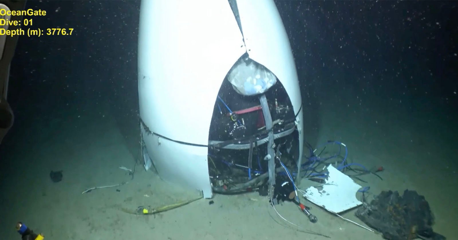 A damaged submersible capsule rests on the ocean floor. The capsule's outer shell is cracked open, revealing its internal components and various cables. The depth is shown as 3776.7 meters. The text reads "OceanGate Dive: 01".